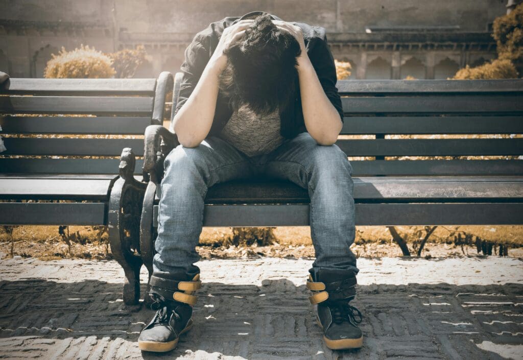 A person is sitting on a bench, leaning forward with their head in their hands, appearing distressed or upset.
