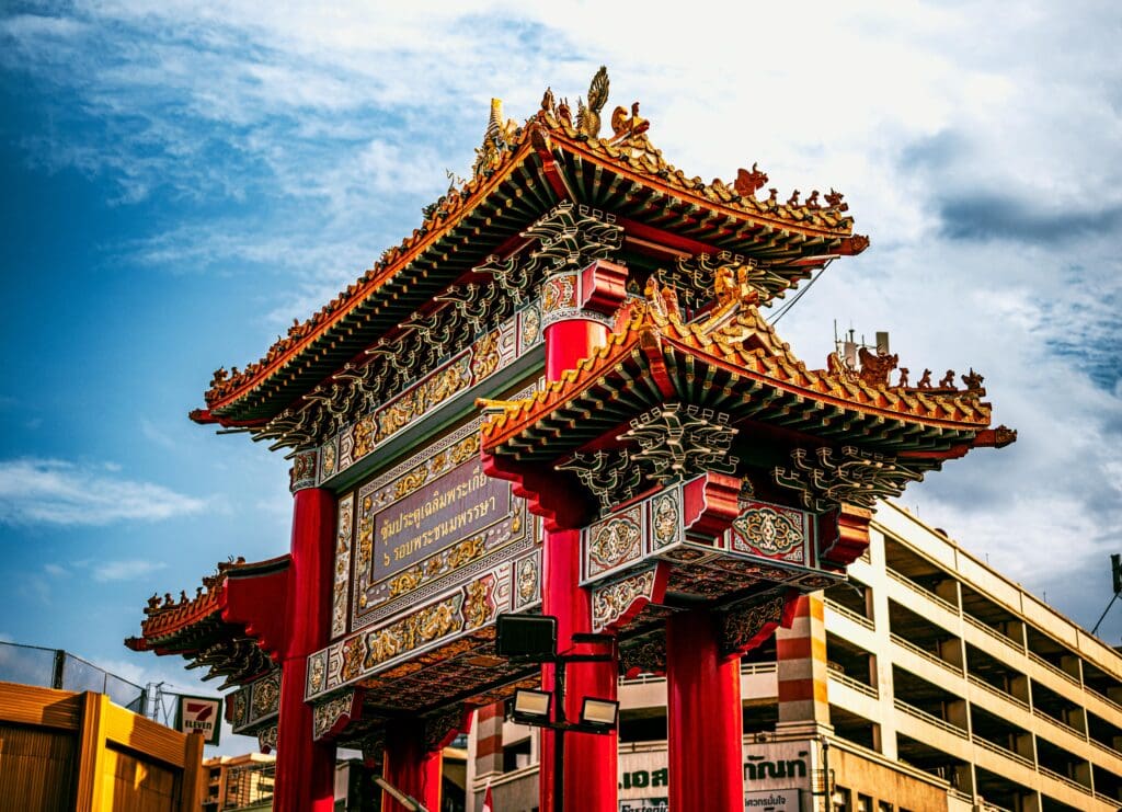 Ornate red and gold Chinese-style archway with intricate carvings and a sign with Thai script, set against a blue sky and urban buildings.