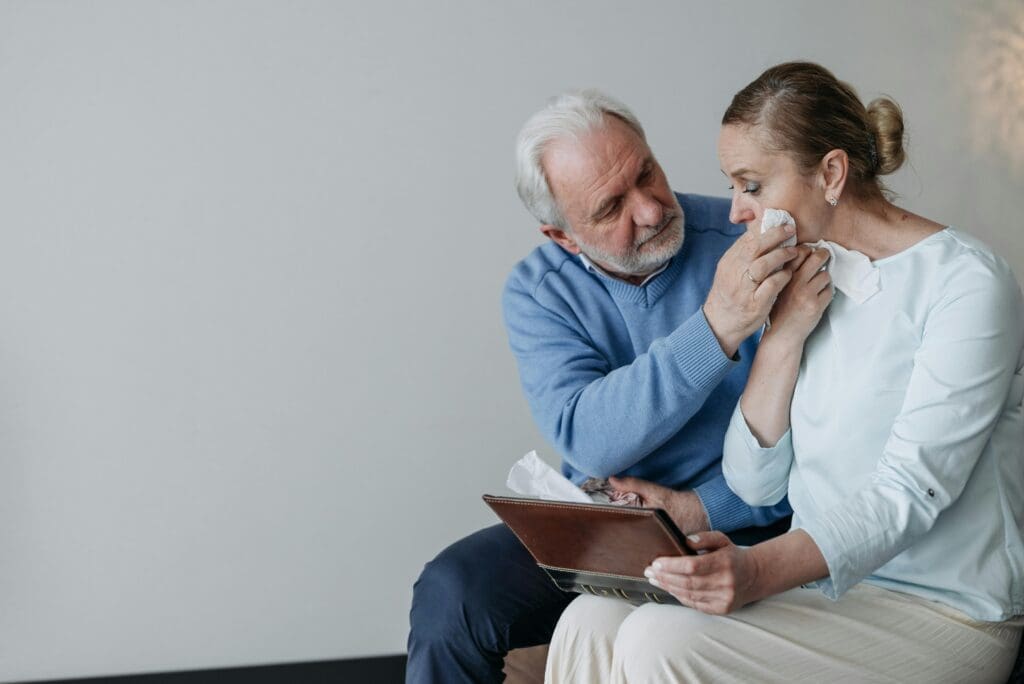 An older man comforts a woman by wiping her tears as she holds a tablet, both sitting together indoors.