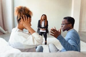 A couple sits on a couch having a tense discussion while a therapist observes and takes notes during a counseling session.