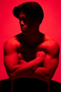 Shirtless man with muscular arms sits with his arms crossed over a chair, lit by red lighting, looking to the side.