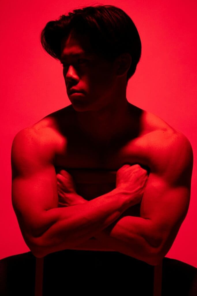 Shirtless man with muscular arms sits with his arms crossed over a chair, lit by red lighting, looking to the side.
