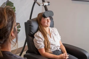 A woman sits in a recliner with her eyes closed while a medical device is positioned over her forehead; another person is seated nearby.