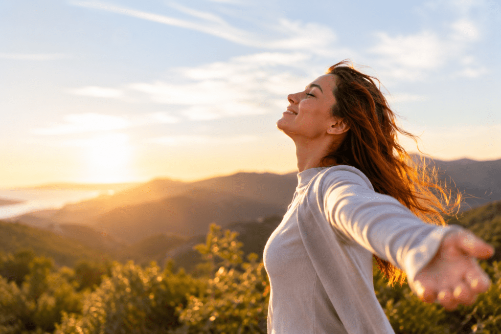Woman standing outdoors on a hillside at sunset with arms outstretched, eyes closed, and a relaxed expression, with a scenic landscape in the background.
