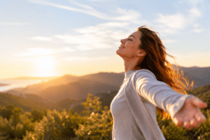 Woman standing outdoors on a hillside at sunset with arms outstretched, eyes closed, and a relaxed expression, with a scenic landscape in the background.