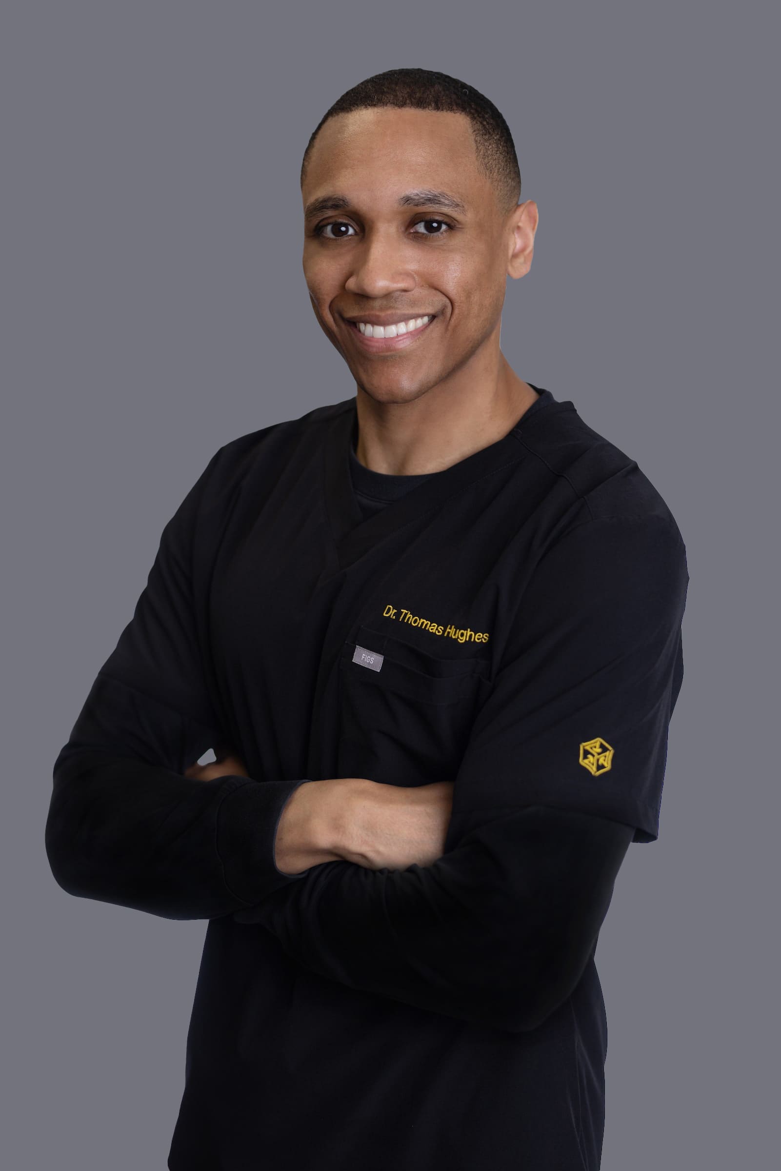 A man wearing black medical scrubs with “Dr. Thomas Hughes” embroidered on the chest stands smiling with arms crossed against a plain gray background.