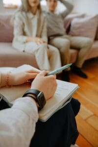 Close-up of a person writing in a notebook with two people sitting on a couch in the background, suggesting a therapy or counseling session.