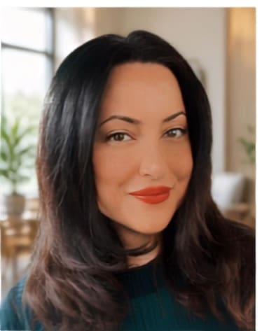 Woman with long dark hair and red lipstick smiling slightly, wearing a dark top, shown indoors with blurred plants and light-colored furniture in the background.