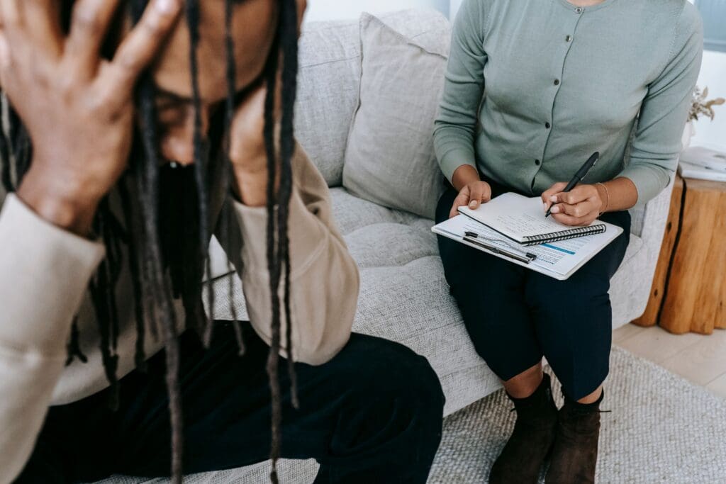 A person holds their head in distress while sitting on a couch, and another person next to them takes notes on a clipboard, suggesting a counseling or therapy session.