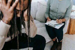 A person holds their head in distress while sitting on a couch, and another person next to them takes notes on a clipboard, suggesting a counseling or therapy session.