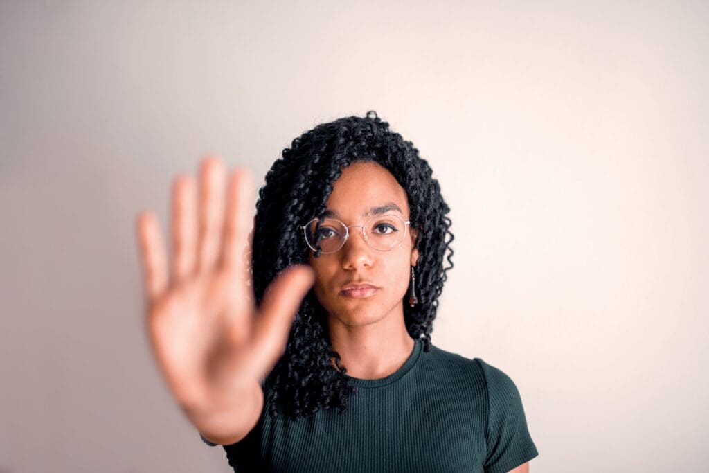 Person with curly hair and glasses holds up hand in a stopping gesture, facing the camera against a plain, light-colored background.