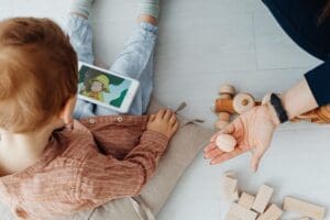 A young child sits on the floor watching a cartoon on a smartphone, while an adult shows a seashell and wooden toys are scattered nearby.