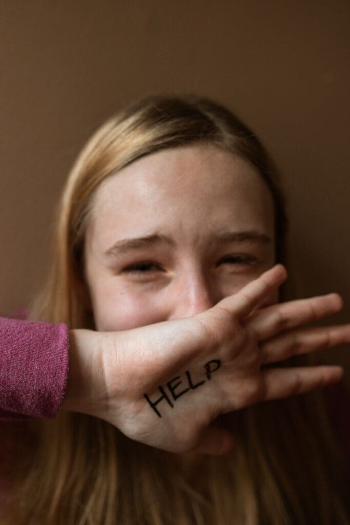 A girl with light brown hair holds her hand over her mouth, showing the word “HELP” written on her palm.