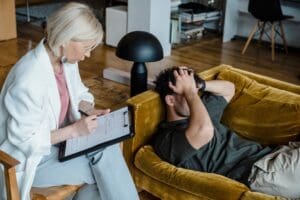 A woman holding a clipboard sits beside a man lying on a couch with his face in his hands in a modern living room.